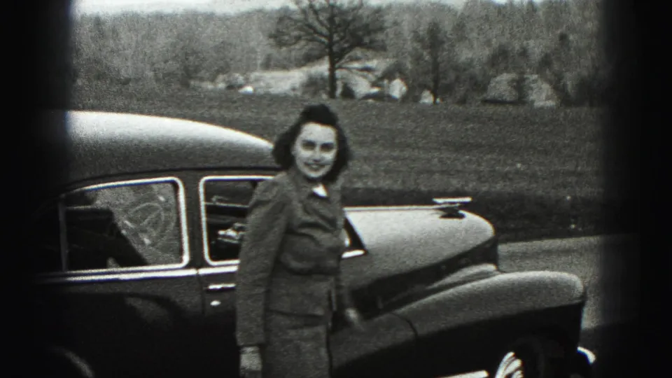 In 1946 New Cumberland, Pennsylvania, a woman in a tailored suit faces the camera beside a rounded dark car in an outdoor setting.