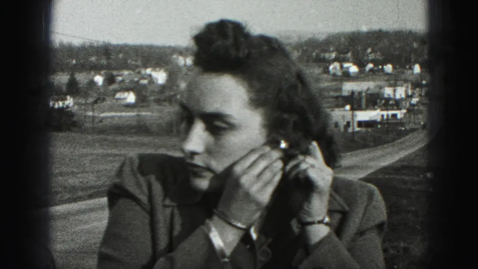 A close 8mm frame shows the woman adjusting an earring, with bracelets visible and a New Cumberland hillside settlement in the background.