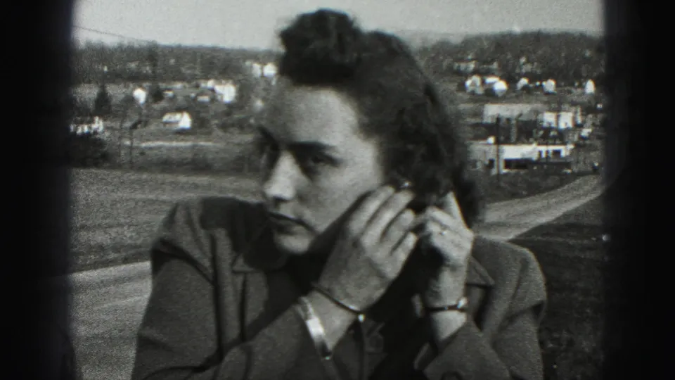 The woman looks aside while adjusting her earring, framed close against roads, houses, and low hills in 1946 Pennsylvania footage.