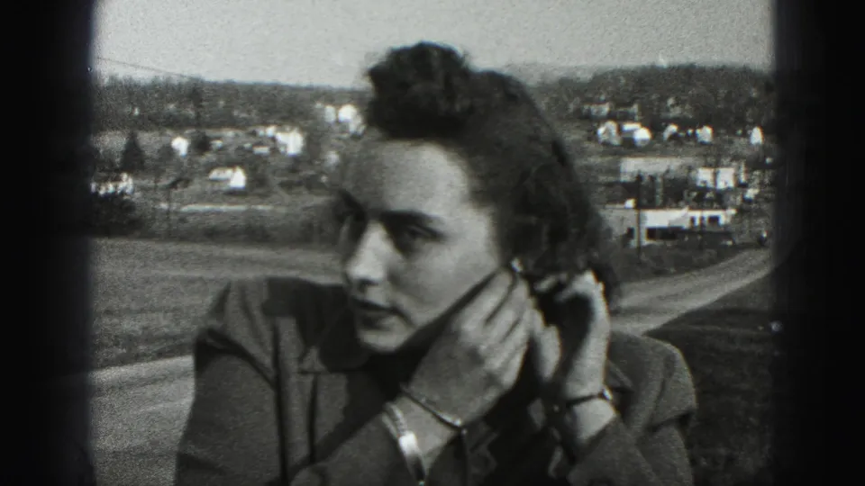 In 1946 New Cumberland, Pennsylvania footage, a woman in a tailored jacket adjusts her left earring with a small town visible beyond.