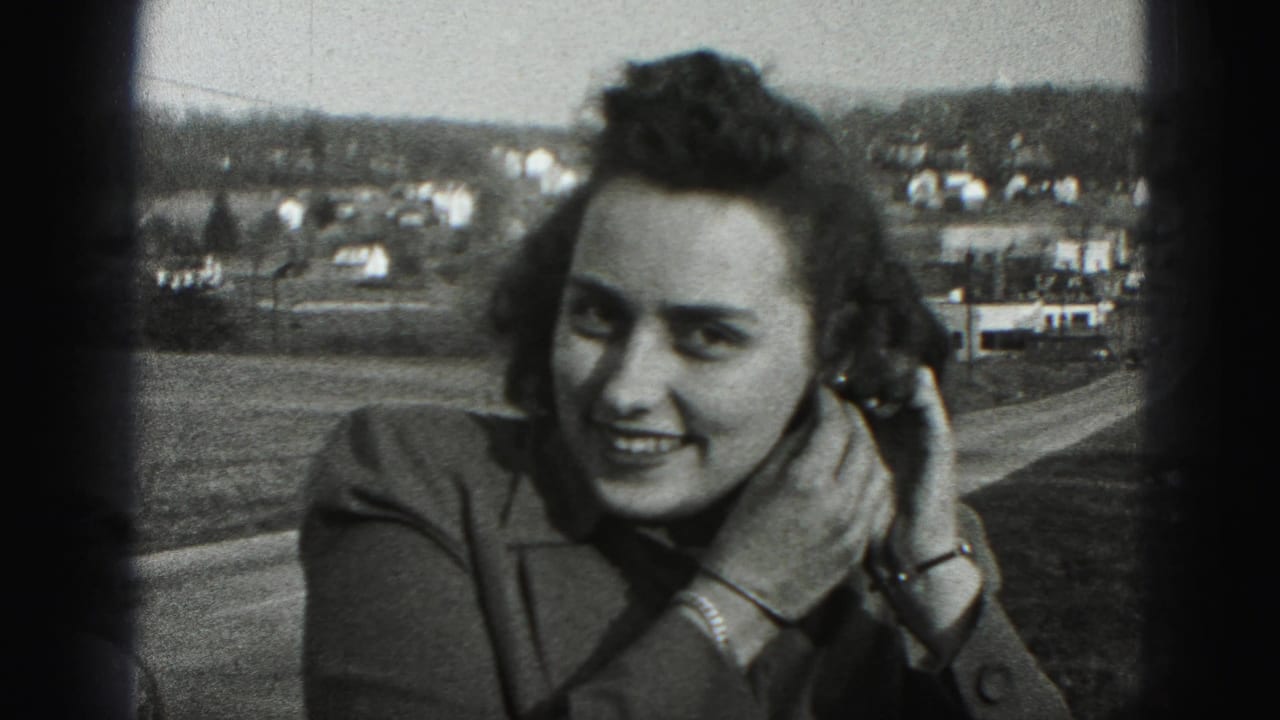 In the 1946 home-movie frame, the woman smiles toward the camera as she continues adjusting her left earring near a town view.