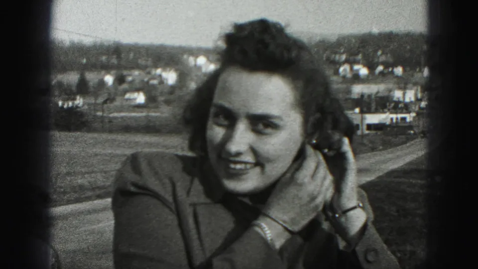 In the 1946 home-movie frame, the woman smiles toward the camera as she continues adjusting her left earring near a town view.