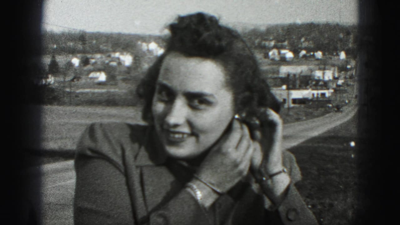 A smiling woman in 1946 New Cumberland footage touches her left earring, with dark frame edges and town buildings visible behind her.