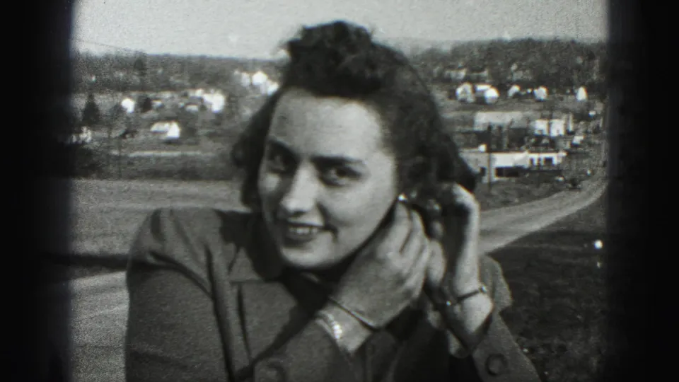 A smiling woman in 1946 New Cumberland footage touches her left earring, with dark frame edges and town buildings visible behind her.