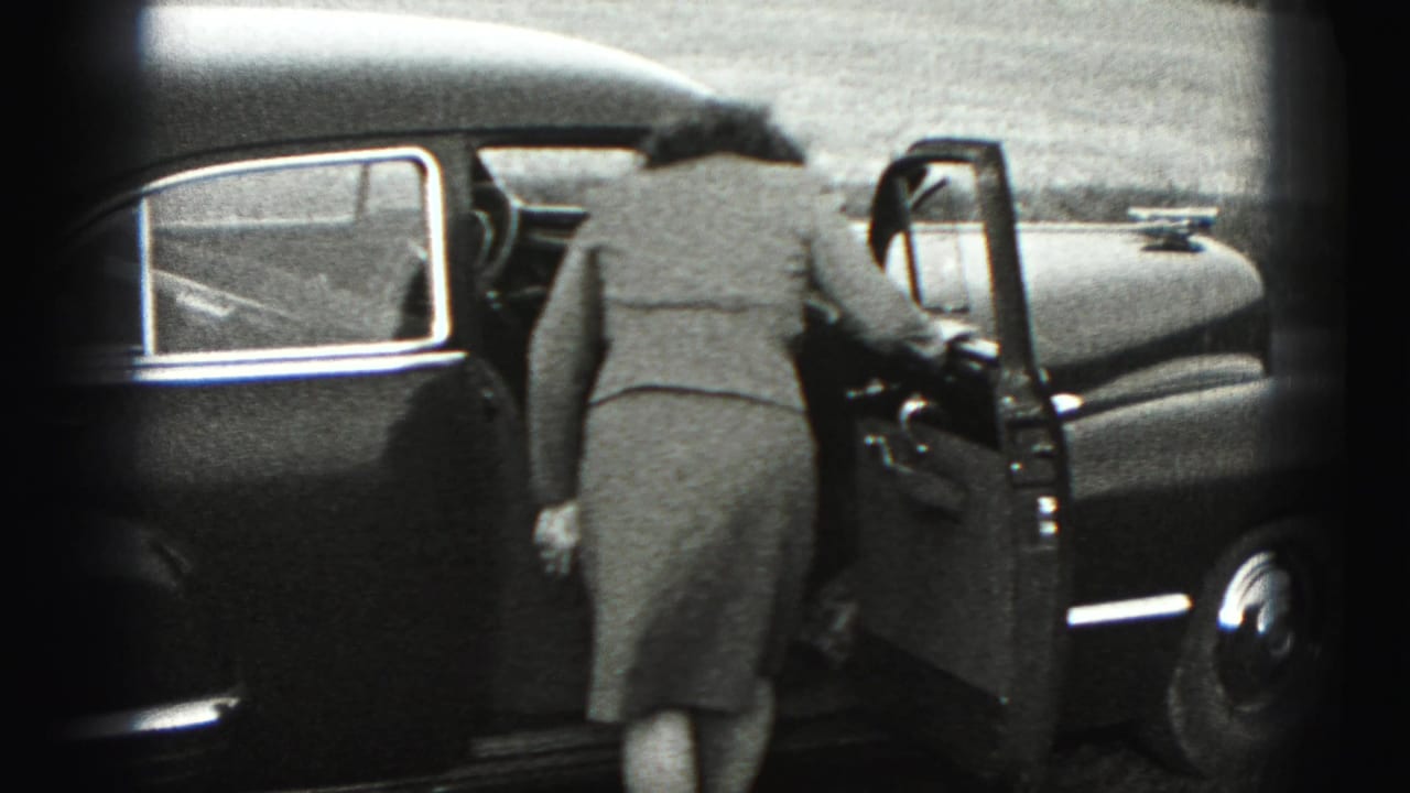 The woman opens the driver-side door of the dark sedan and begins to lean inside in the 1946 home-movie scene.