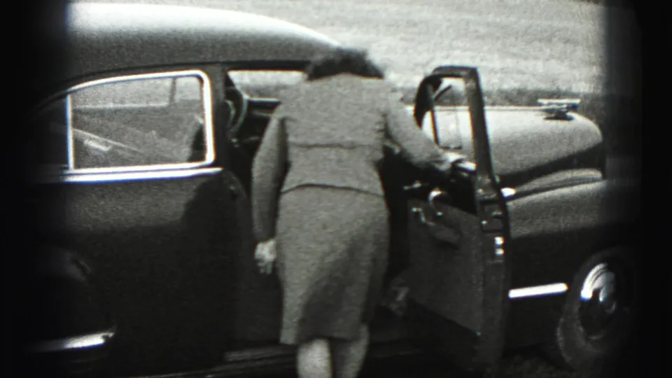 The woman opens the driver-side door of the dark sedan and begins to lean inside in the 1946 home-movie scene.