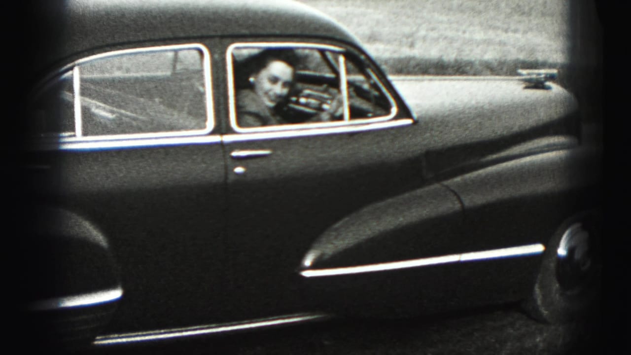 The woman smiles and waves from inside the driver's seat of the dark sedan.