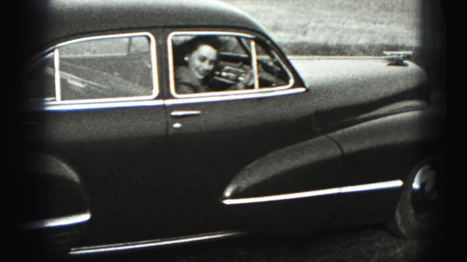 The woman smiles and waves from inside the driver's seat of the dark sedan.