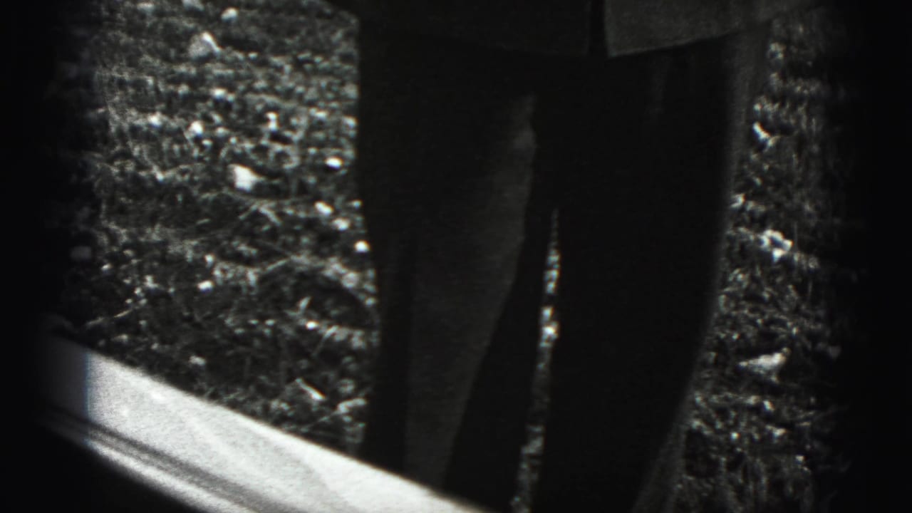 Lower legs of a standing person seen beyond a bright frame edge on gravel.