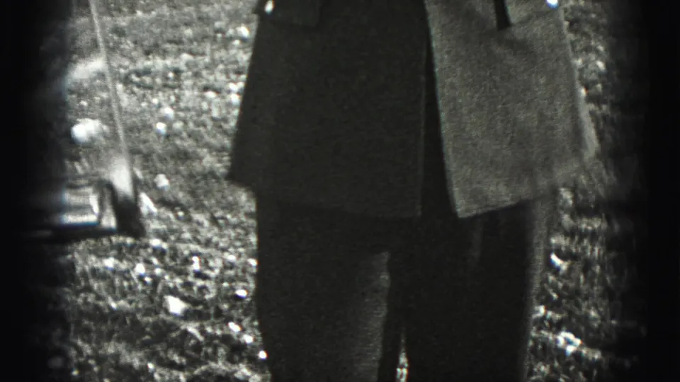 The camera catches part of the returned soldier's lower jacket and legs beside a dark edge, with gravel and yard detail behind.