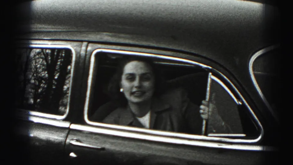 A woman seated inside a mid-century car faces the camera through the side window in New Cumberland home-movie footage.