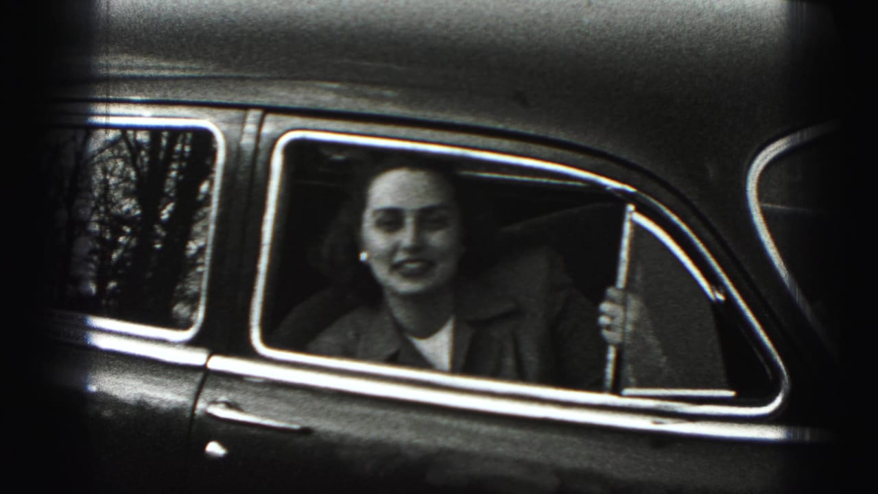 The woman looks toward the camera from the backseat, holding the open window frame of a 1940s car.