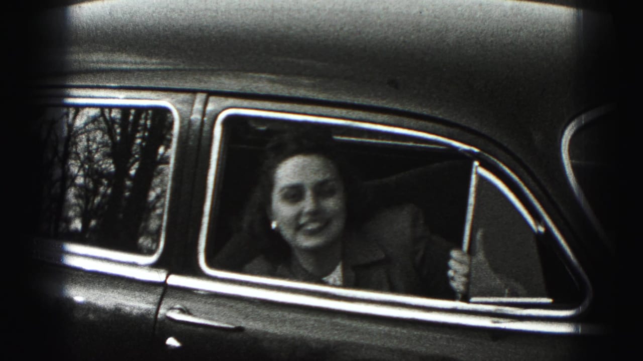 The woman smiles wider from the open rear window of the vintage car, with soft 8mm grain and edge shadow visible.