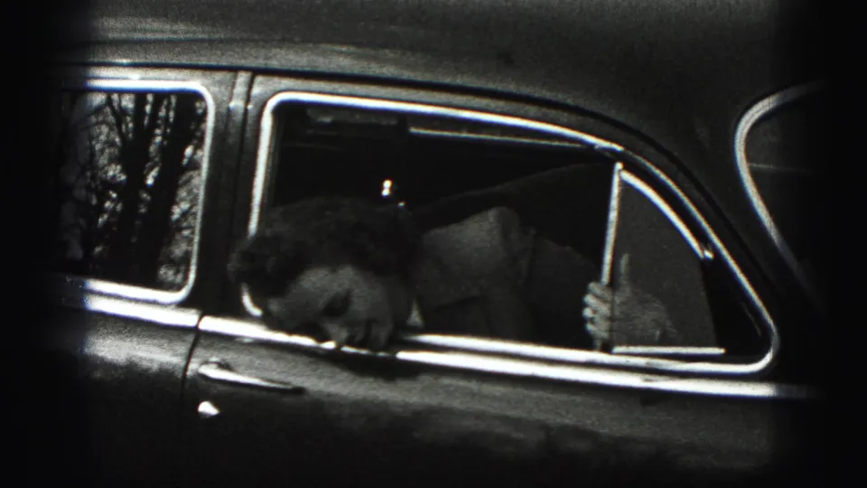A woman bends downward inside the rear passenger window of a 1940s car, with trees reflected in the glass.