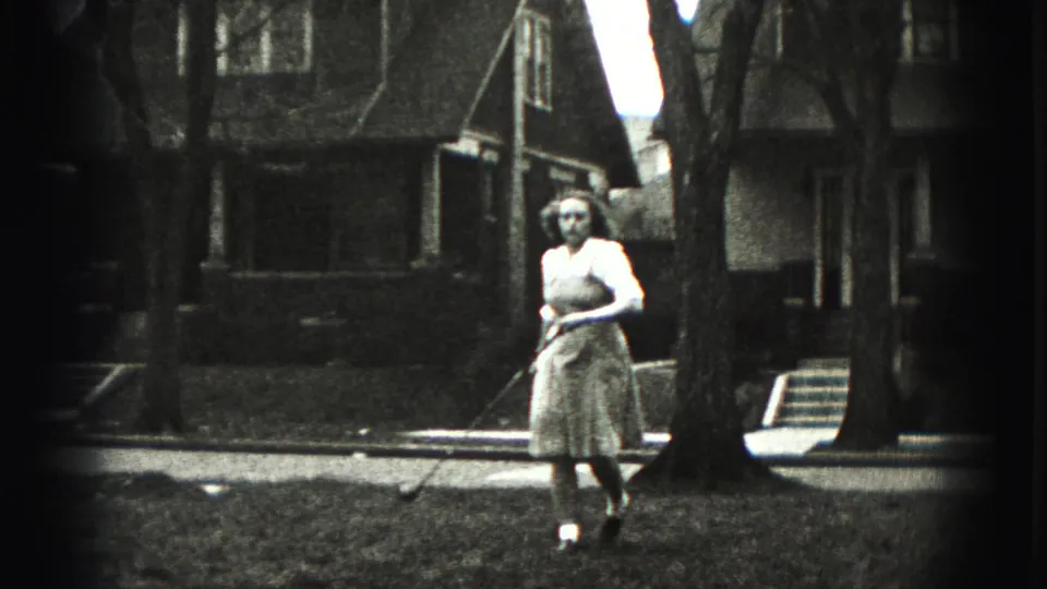 A woman in a light dress walks across a residential lawn with a golf club, framed by houses and tree trunks.