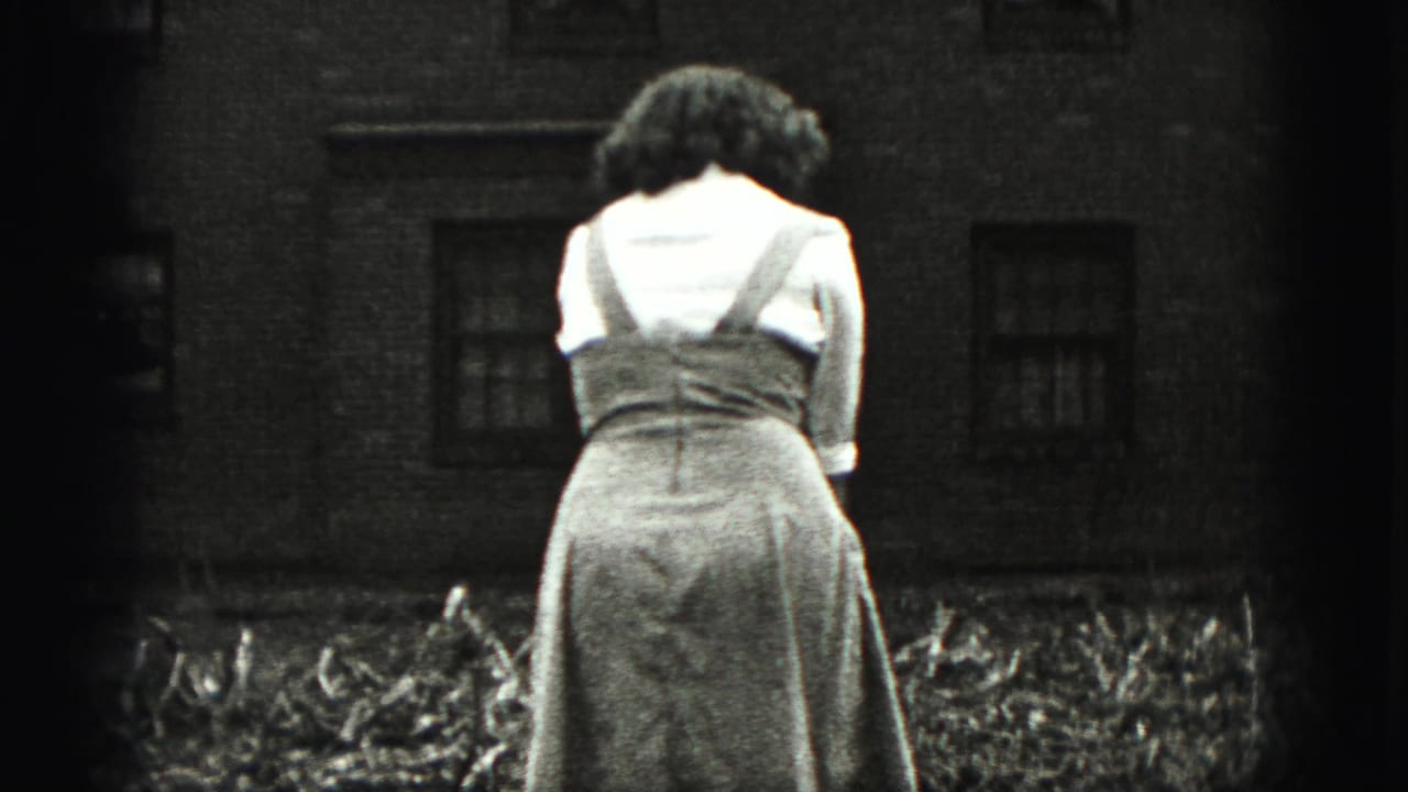 A woman in a light blouse and dress stands outdoors in the New Cumberland yard, with a brick house and garden debris behind her.