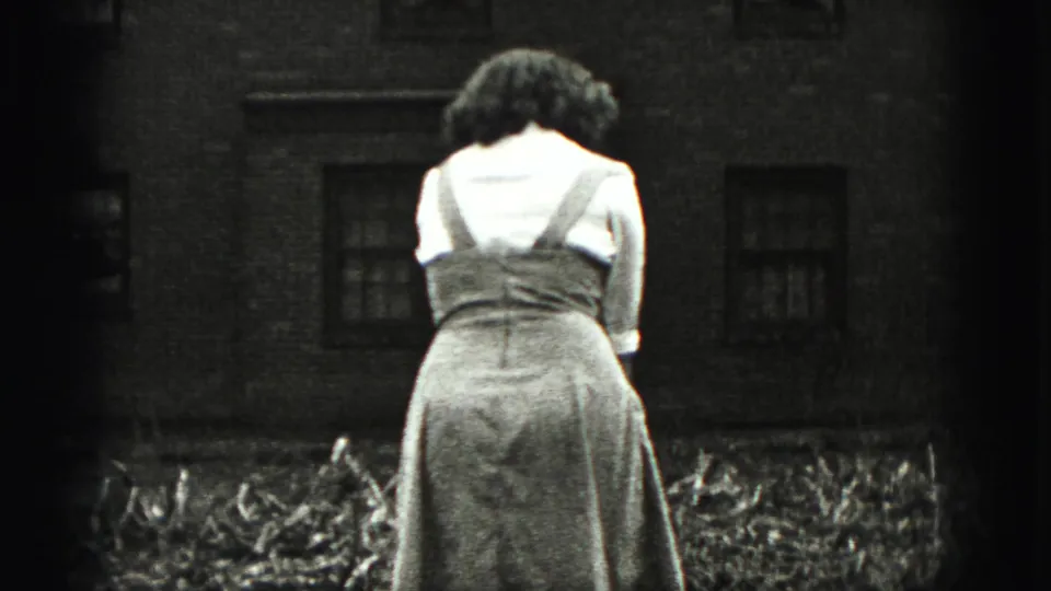 A woman in a light blouse and dress stands outdoors in the New Cumberland yard, with a brick house and garden debris behind her.