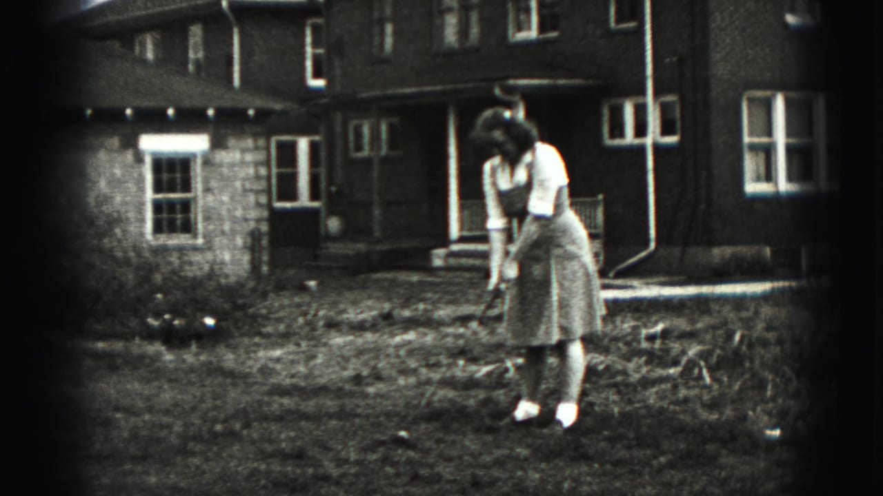 Woman stands in a yard addressing a golf ball with a club in front of a brick house.