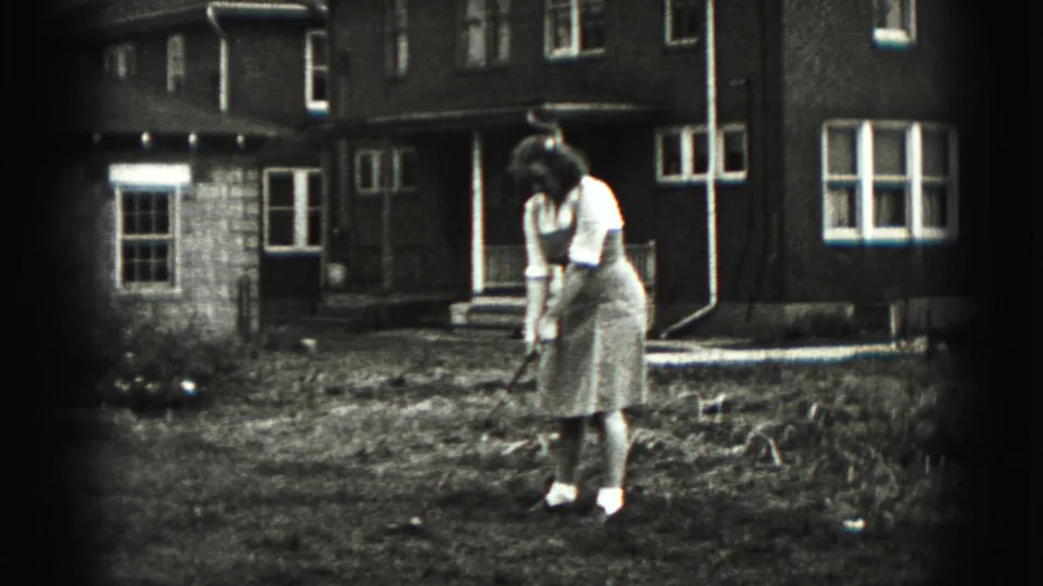 A woman in a mid-1940s domestic yard stance looks down toward the ground while holding a golf club near a brick house.