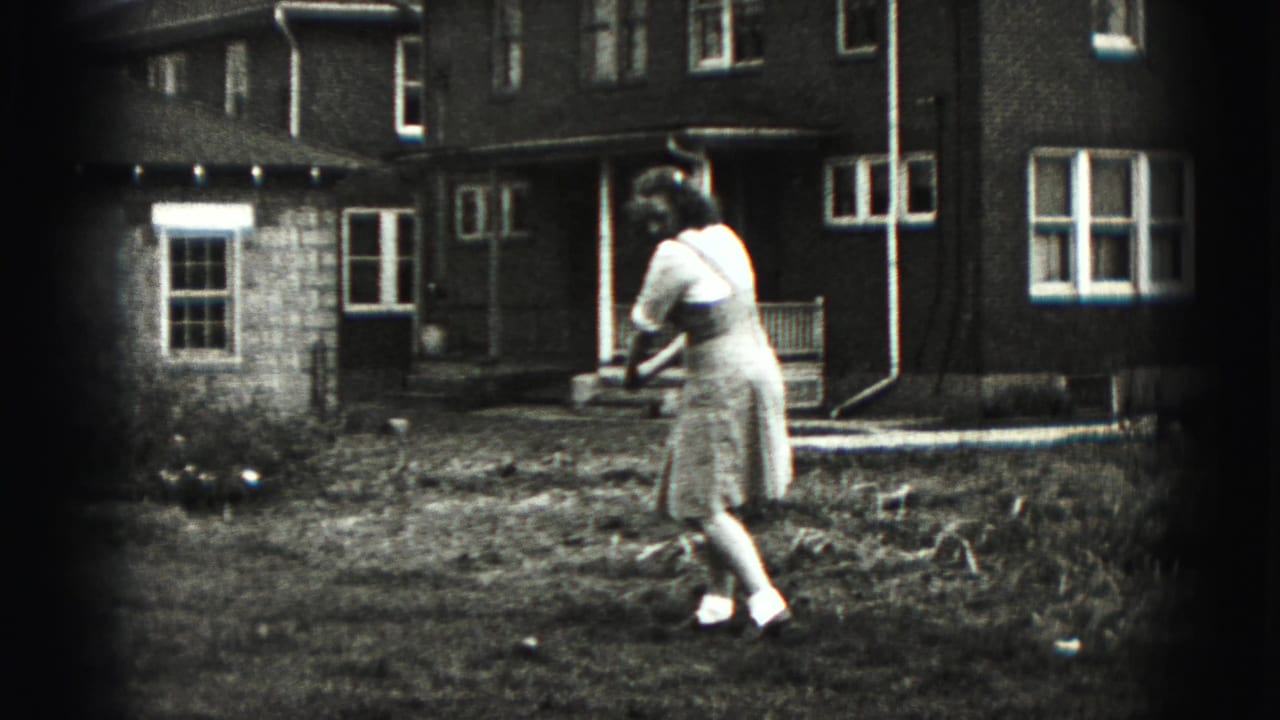 The woman starts rotating into a backyard golf swing, filmed in black-and-white 8mm against a New Cumberland house exterior.