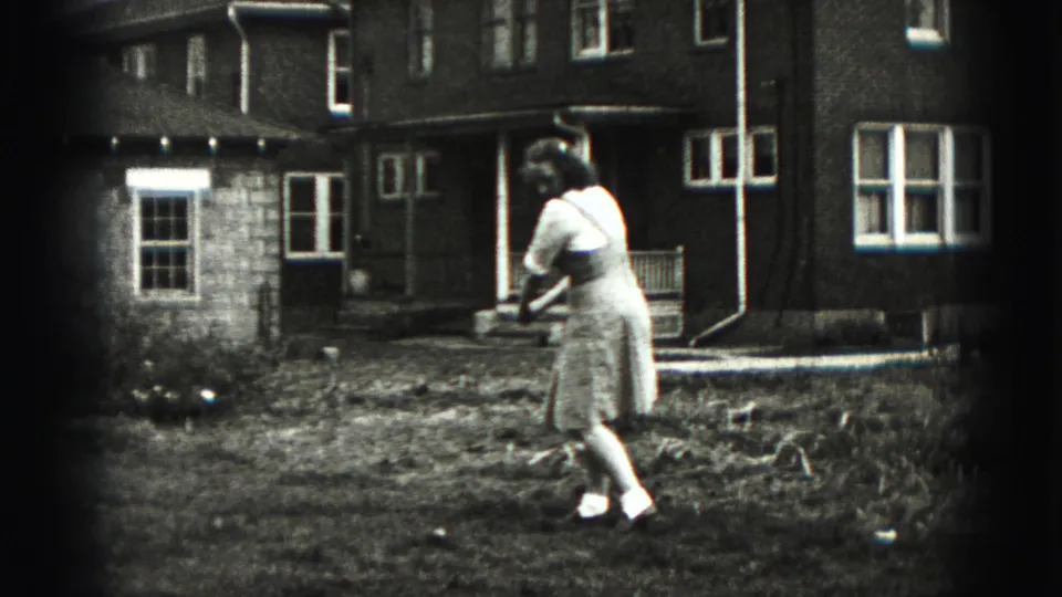 The woman starts rotating into a backyard golf swing, filmed in black-and-white 8mm against a New Cumberland house exterior.
