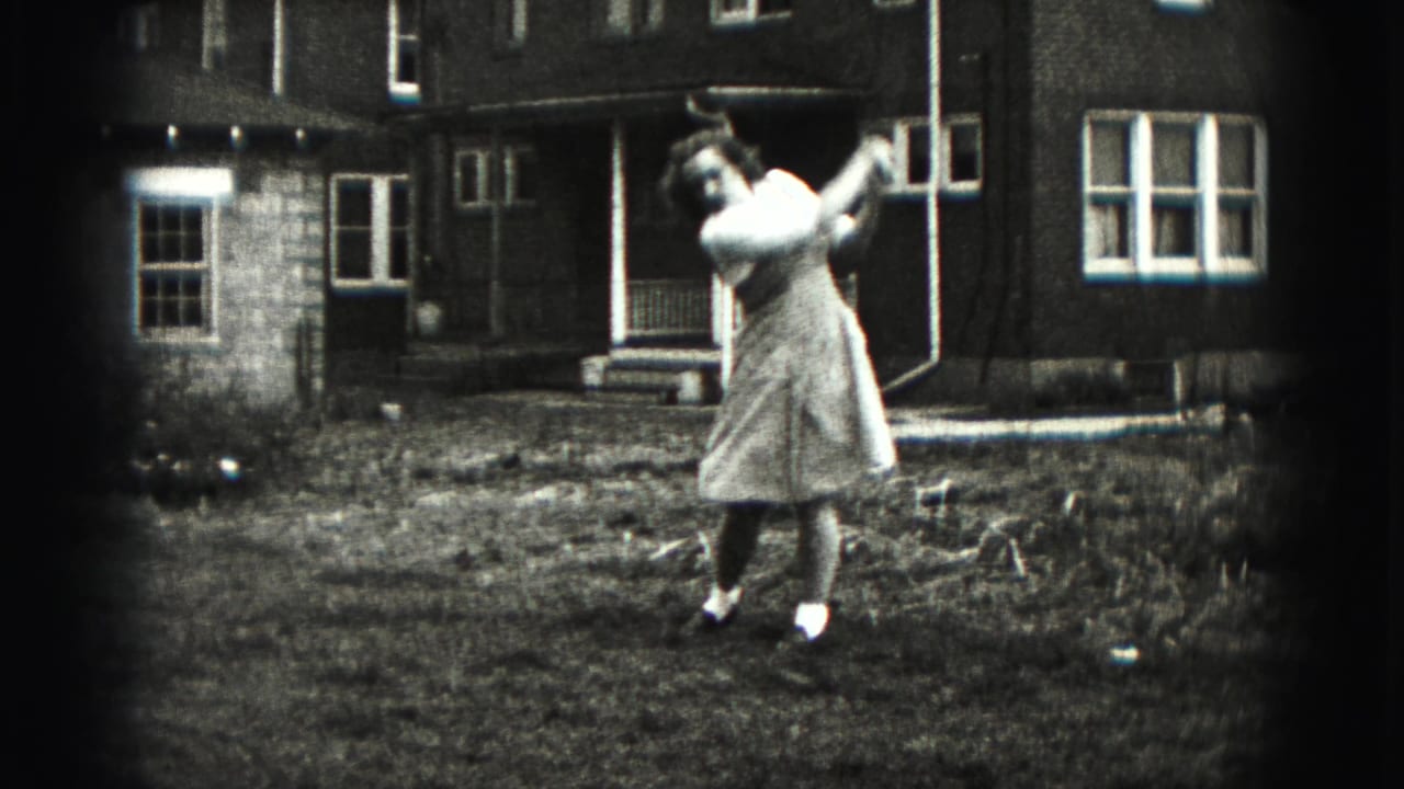 A woman in a light dress completes her backyard golf swing, with residential windows and porch details visible behind her.