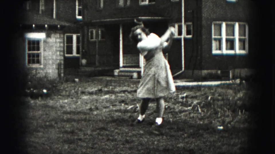 A woman in a light dress completes her backyard golf swing, with residential windows and porch details visible behind her.