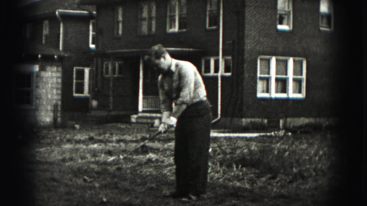 Man bends over a golf club in a yard in front of a brick house.