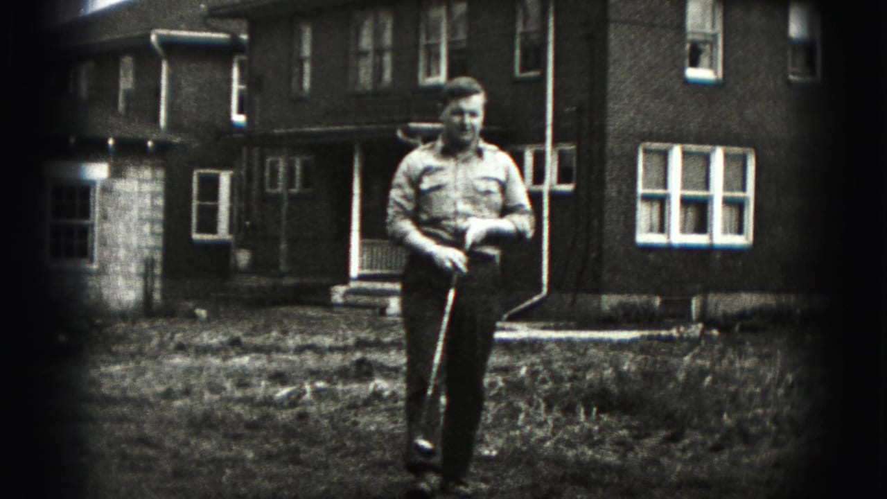 A man in a button-up shirt walks through a New Cumberland front yard carrying a golf club, with brick homes behind him.