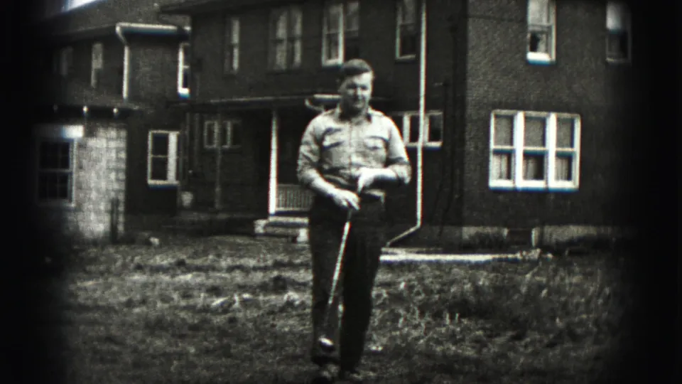 A man in a button-up shirt walks through a New Cumberland front yard carrying a golf club, with brick homes behind him.