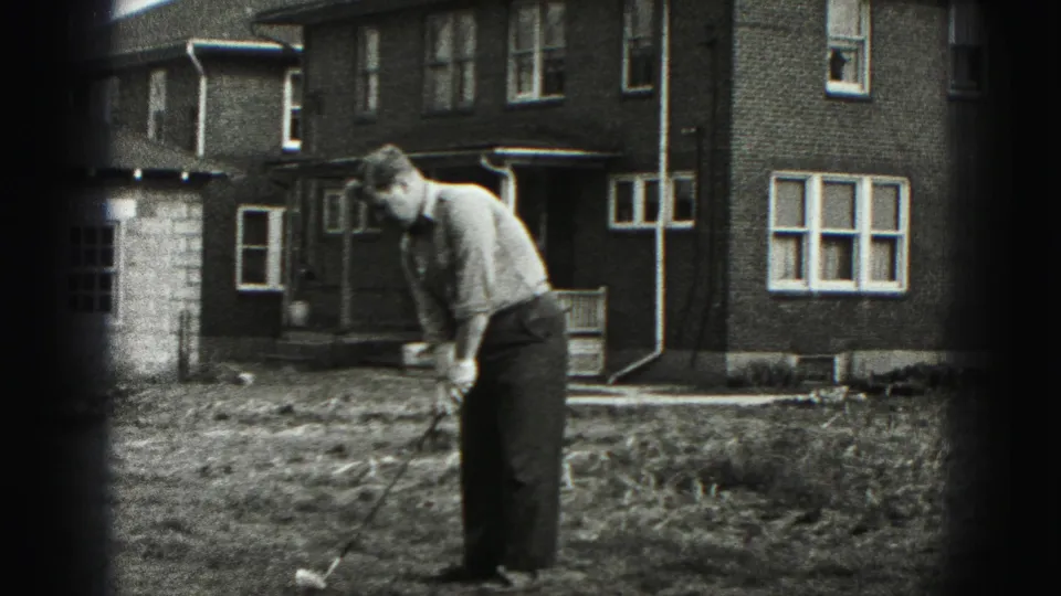 The man remains focused over the ball with a golf club in a New Cumberland yard, framed by brick walls and windows.
