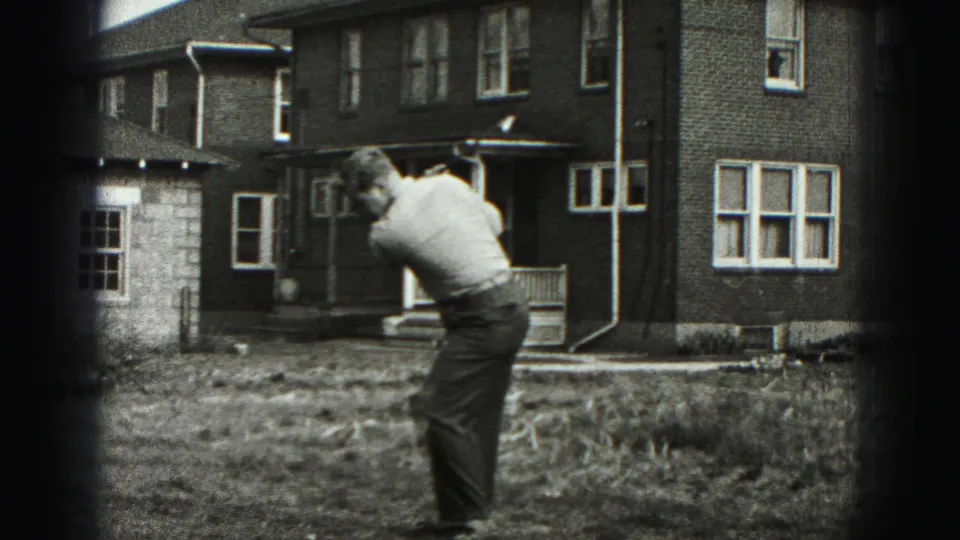 The man follows through on a backyard golf swing in 1946 home-movie footage, with a brick house behind him.