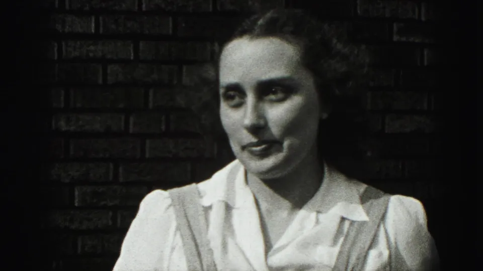 A young woman in a white blouse and suspenders faces the camera against a brick wall in the 1946 New Cumberland home movie.