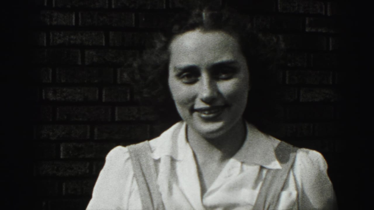 The young woman smiles gently at the camera, her white blouse bright against the dark brick wall in the 1946 footage.