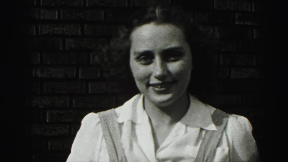 The young woman smiles gently at the camera, her white blouse bright against the dark brick wall in the 1946 footage.