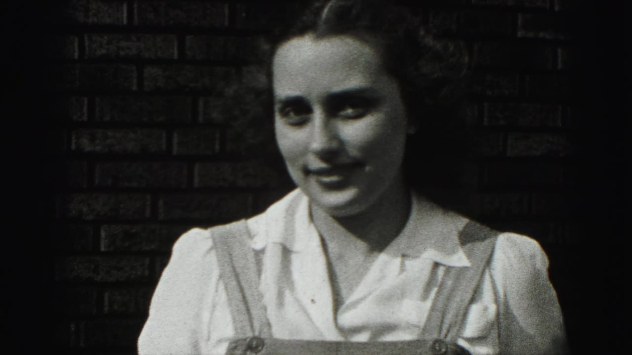 Young woman in a white blouse and straps faces the camera in front of a dark brick wall.