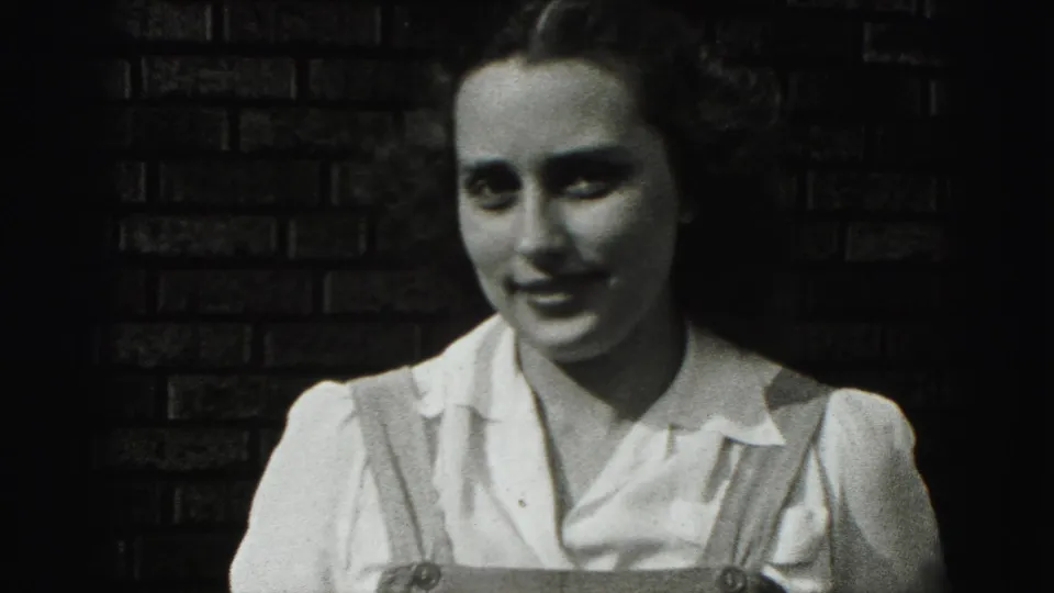 Young woman in 1946 New Cumberland footage faces the camera, wearing a white collared blouse and shoulder straps before a dark brick wall.