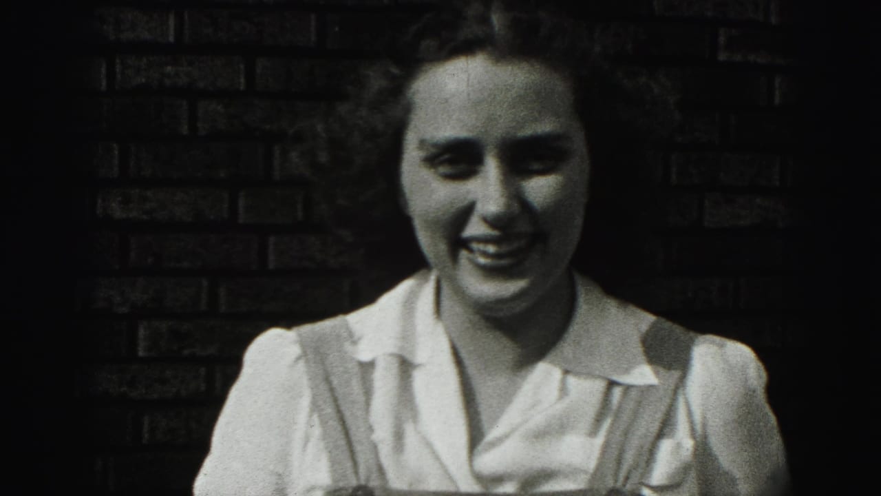 The woman smiles broadly in the 1946 New Cumberland 8mm footage, framed close against a dark brick wall.