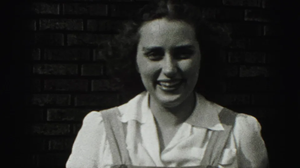 The woman smiles broadly in the 1946 New Cumberland 8mm footage, framed close against a dark brick wall.