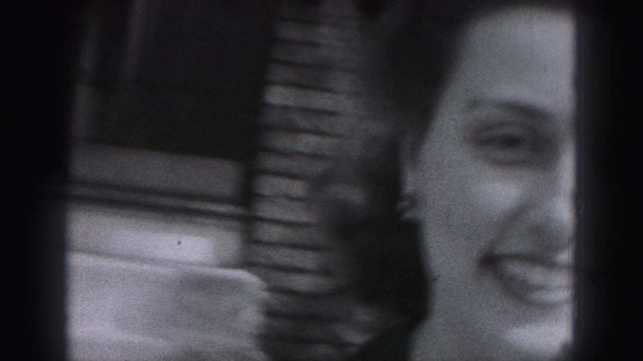 Partial close-up of the smiling woman in soft black-and-white 8mm footage, with brickwork visible behind her.
