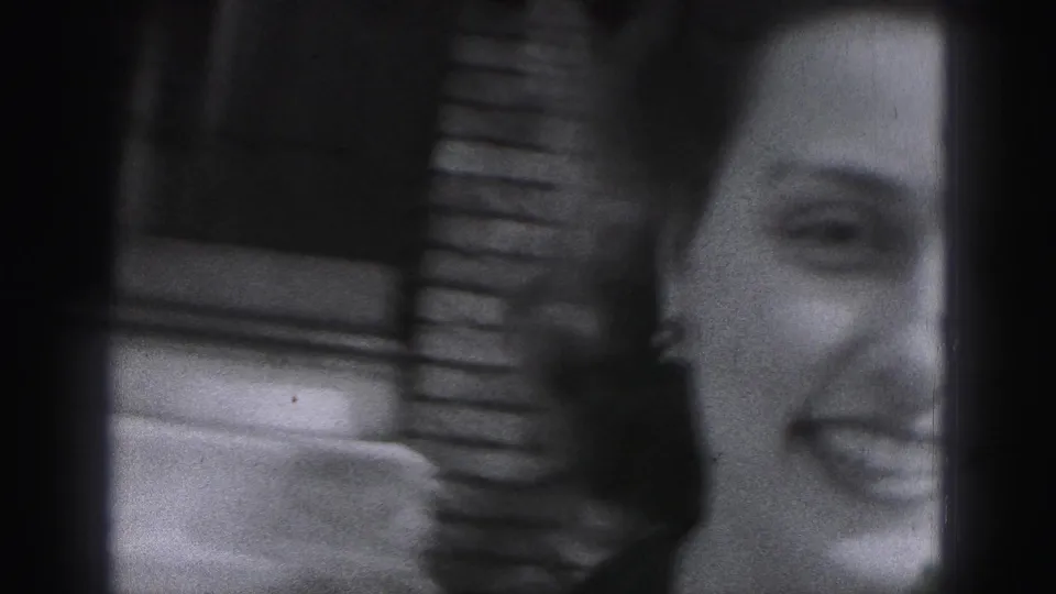 Partial close-up of the smiling woman in soft black-and-white 8mm footage, with brickwork visible behind her.