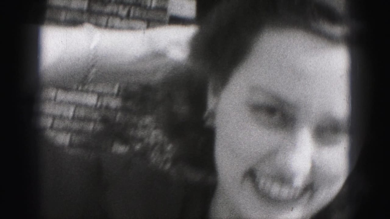 Young woman in a dark dress smiles toward the camera while lifting a dark hat near a brick wall.