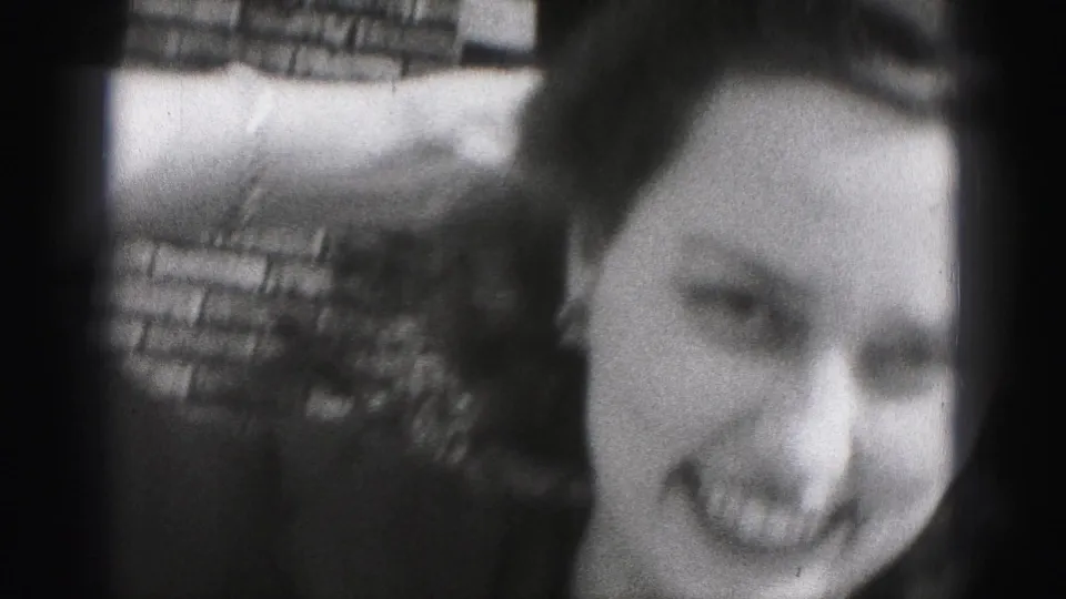 In this 1946 New Cumberland home-movie frame, a young woman in a dark dress smiles as she raises a hat beside a brick wall.