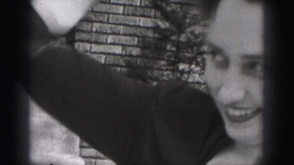 A 1946 home-movie frame partly crops the smiling woman as her dark sleeve and hat pass across the foreground near a brick wall.