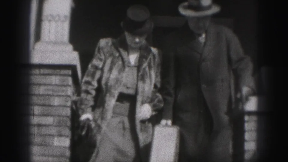 In 1946 New Cumberland home-movie footage, an adult couple in period clothing walks outdoors near steps, the man holding a suitcase.