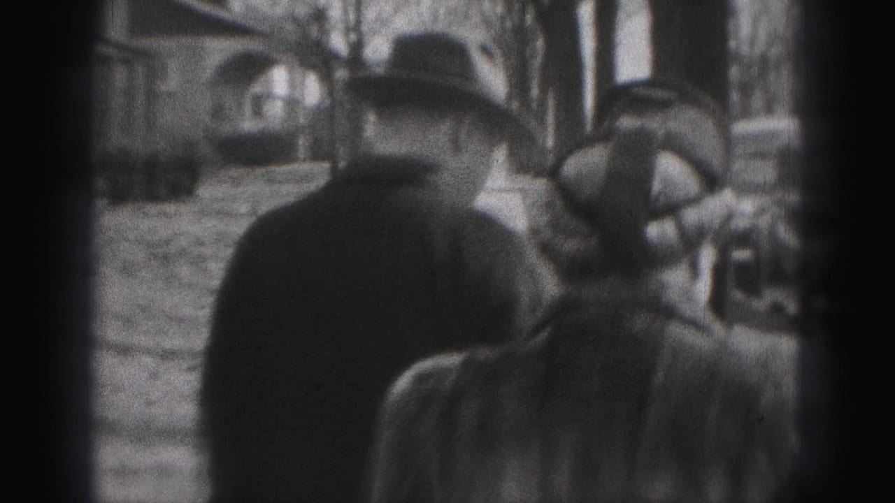 Two adults in coats and hats walking along a sidewalk near parked cars.