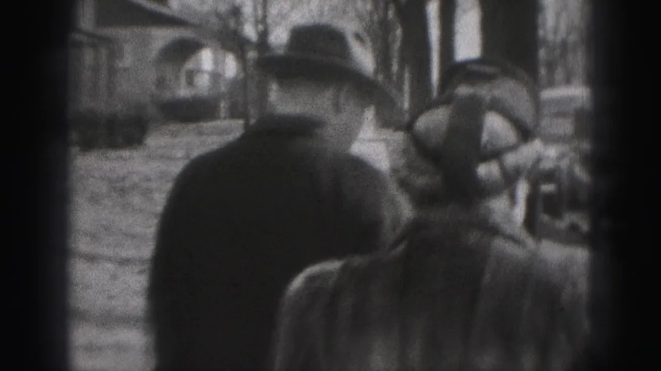 In New Cumberland, Pennsylvania, two adults in 1940s coats and hats walk along a tree-lined sidewalk near parked cars.