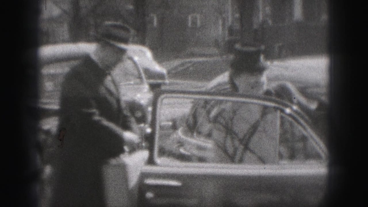 A man in a brimmed hat stands beside a vintage car in New Cumberland, with another adult nearby and reflections in the car glass.