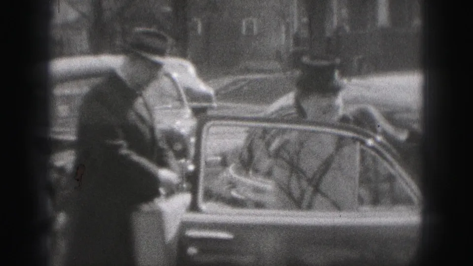 A man in a brimmed hat stands beside a vintage car in New Cumberland, with another adult nearby and reflections in the car glass.