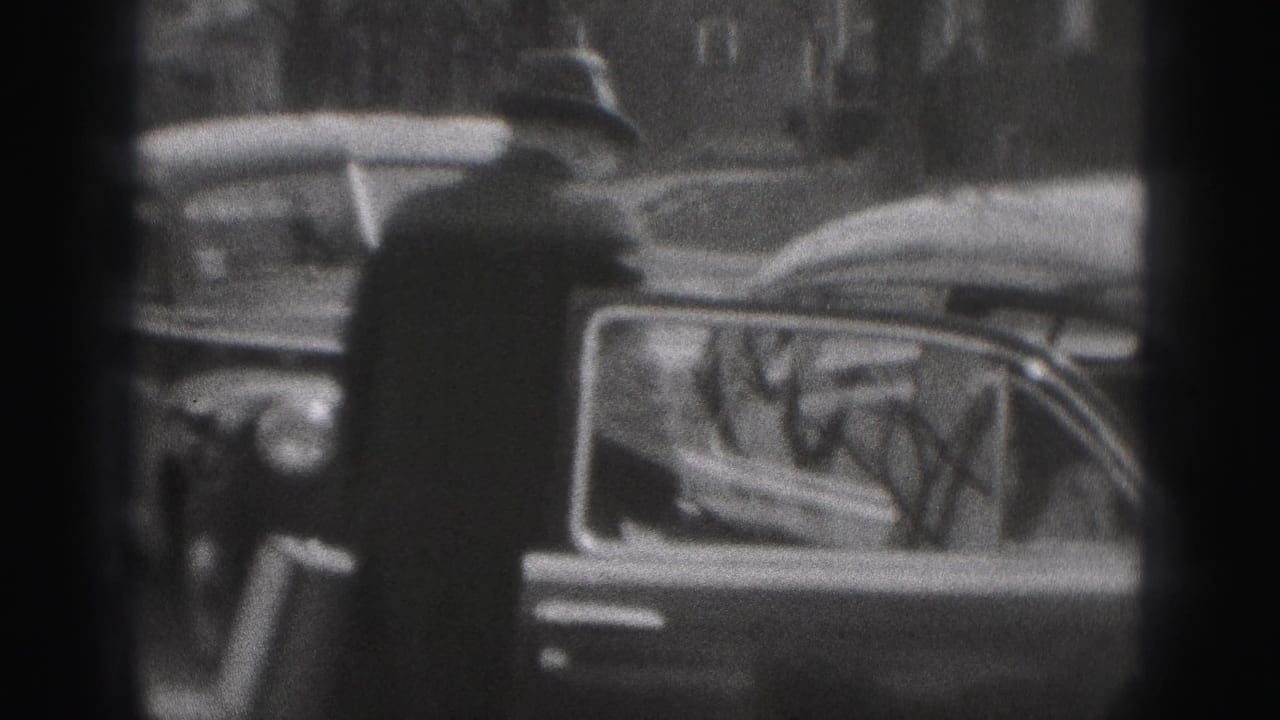 A man in a fedora and dark coat stands near a parked vintage car on the New Cumberland street in black-and-white 8mm footage.